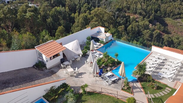 Luchtbeeld van een resort met een zwembad, omringd door ligbedden en parasols, met op de achtergrond een dicht bos en woongebouwen.