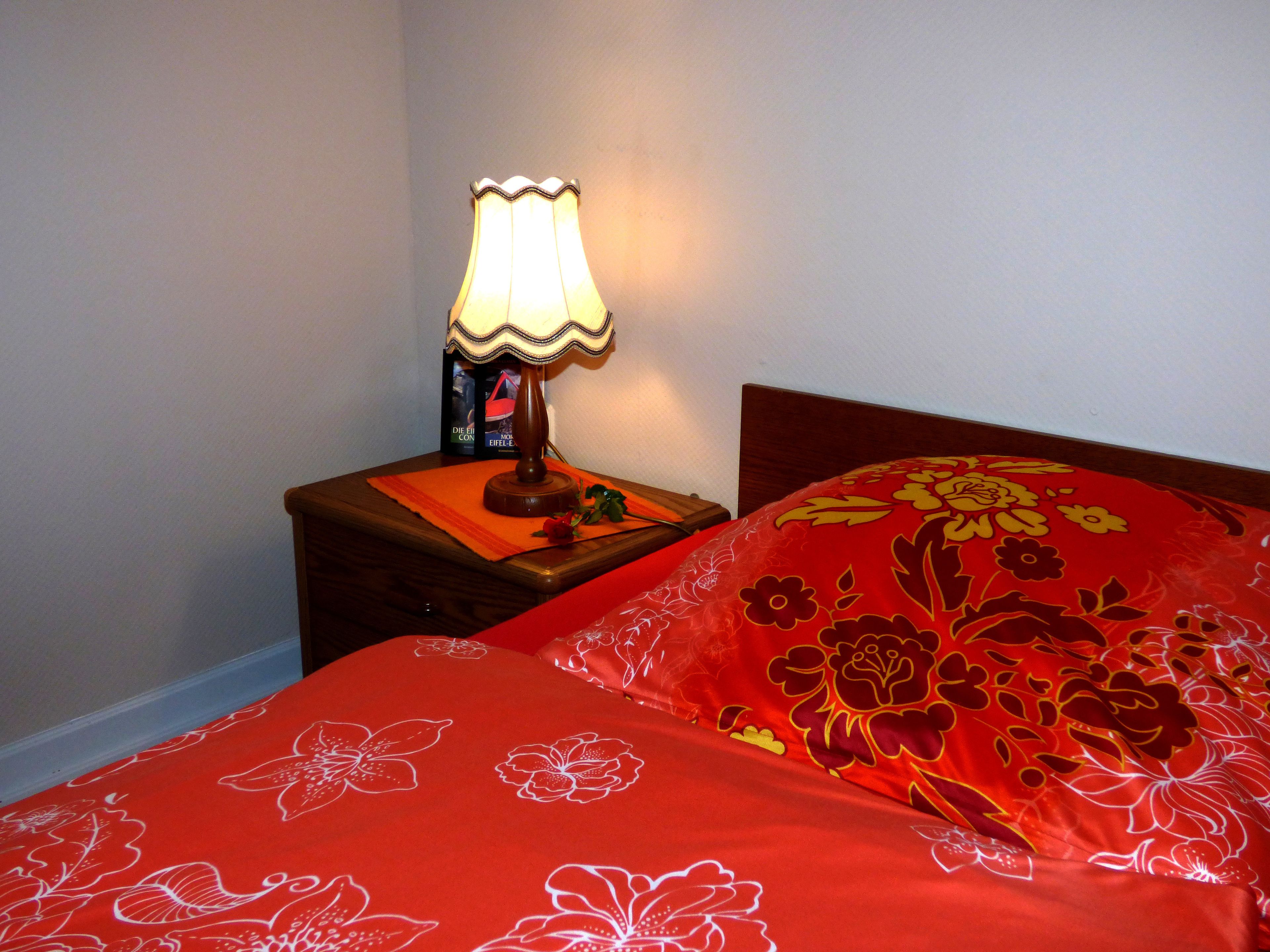 A bed with red floral bedding and a lit table lamp on a wooden nightstand.