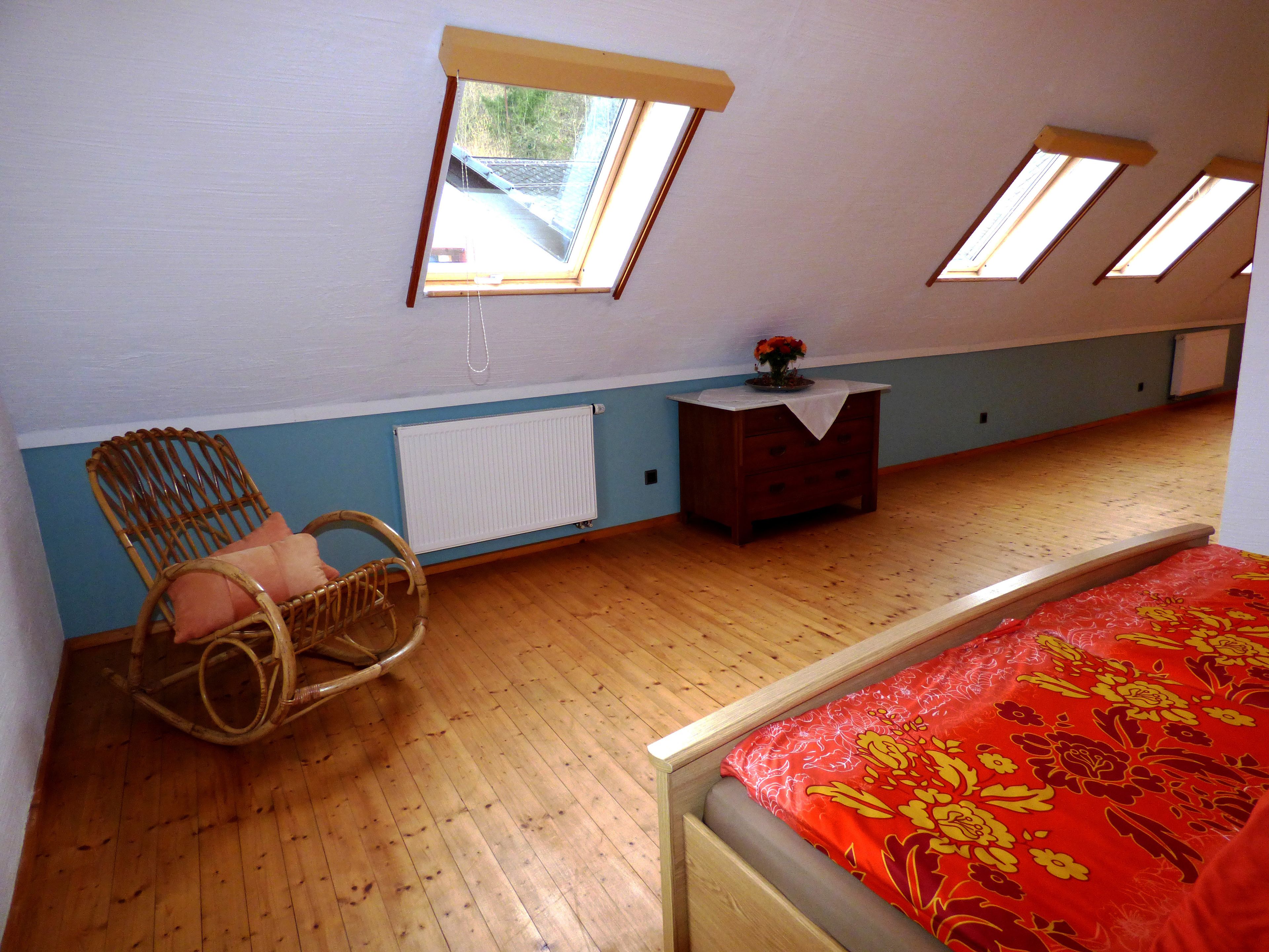 A bedroom with a sloped ceiling, wooden flooring, a bed with a red floral bedspread, a wicker chair, a dresser, and a radiator.