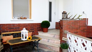 Un petit espace extérieur aménagé avec une table, des chaises et une lanterne. On trouve une plante en pot sur les marches et une pancarte de bienvenue sur le mur.