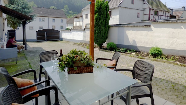 A covered patio with a glass table and chairs, surrounded by a white wall and a garden. - Relaxen im gemütlichen Eifelhaus (Kesseling)
