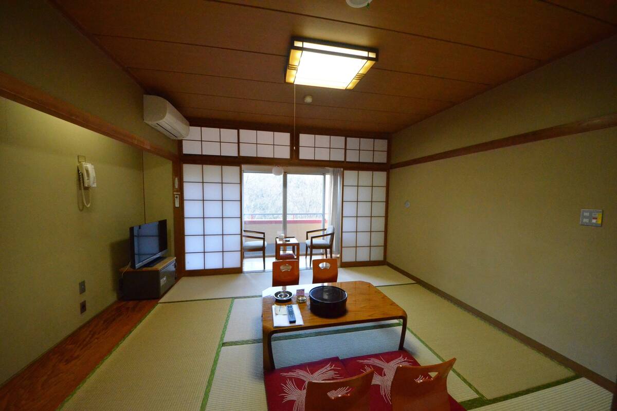 A traditional Japanese room with tatami flooring, a low table with a black bowl, a television, and a sliding door leading to a balcony.