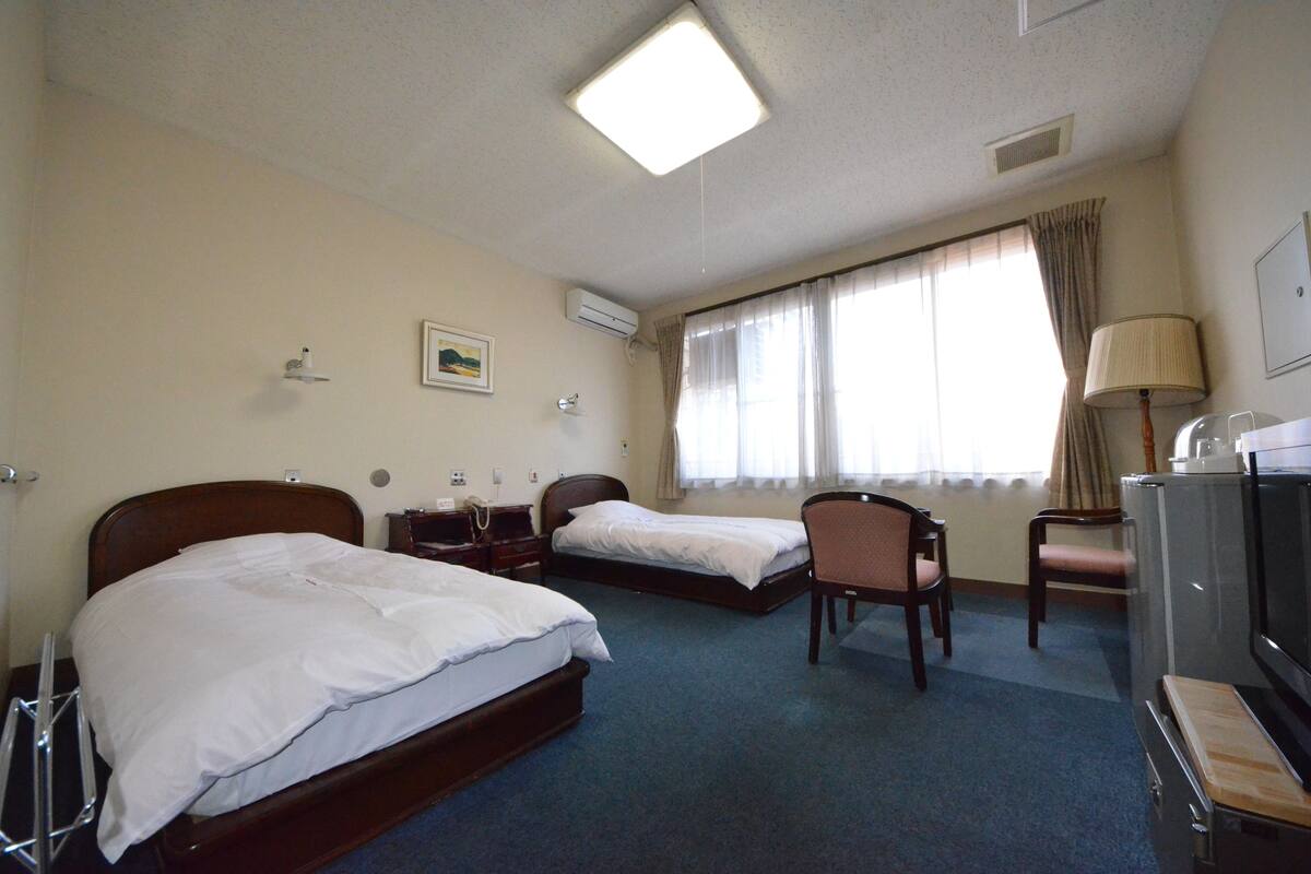 A hotel room with two single beds, a desk with a chair, a television, and a window with curtains.