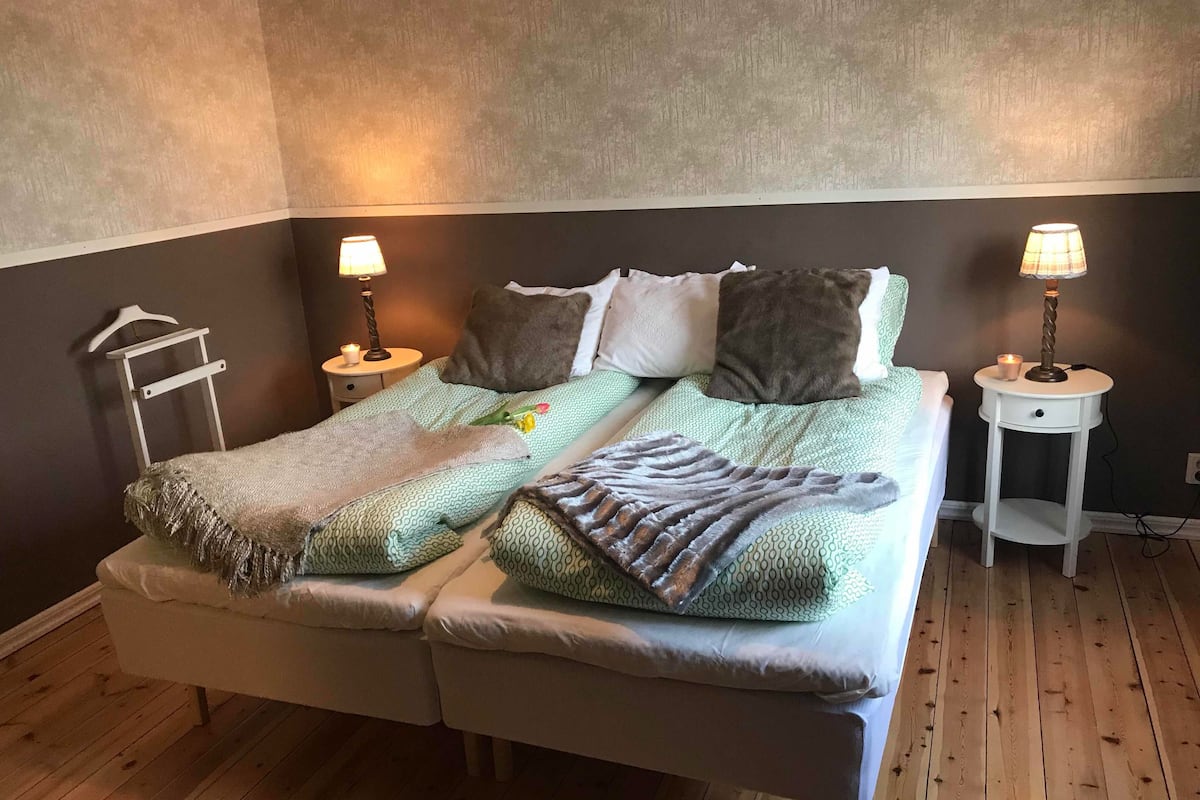 Cama doble con una colcha estampada y dos almohadas, flanqueada por mesitas de noche con lámparas y una vela pequeña.