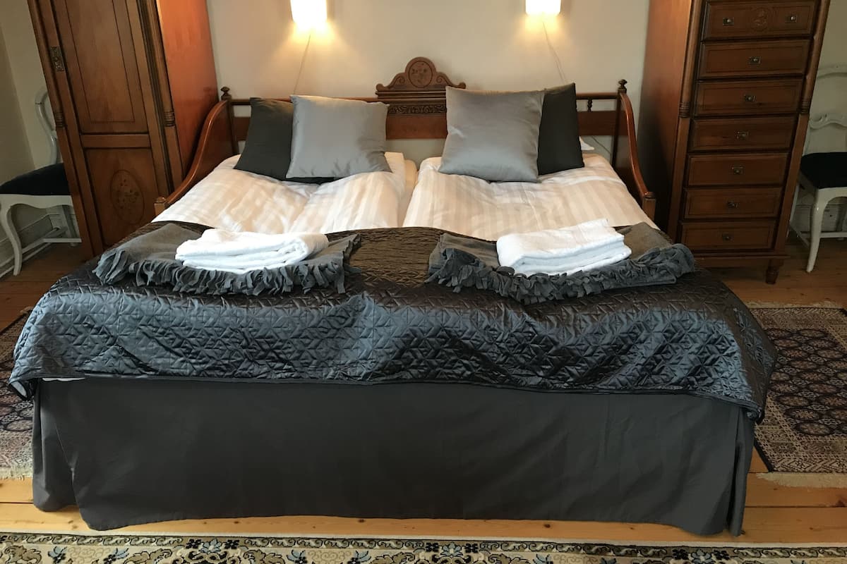 Una cama bien hecha con una colcha oscura y almohadas, un cabecero de madera y dos luces fijadas a la pared.