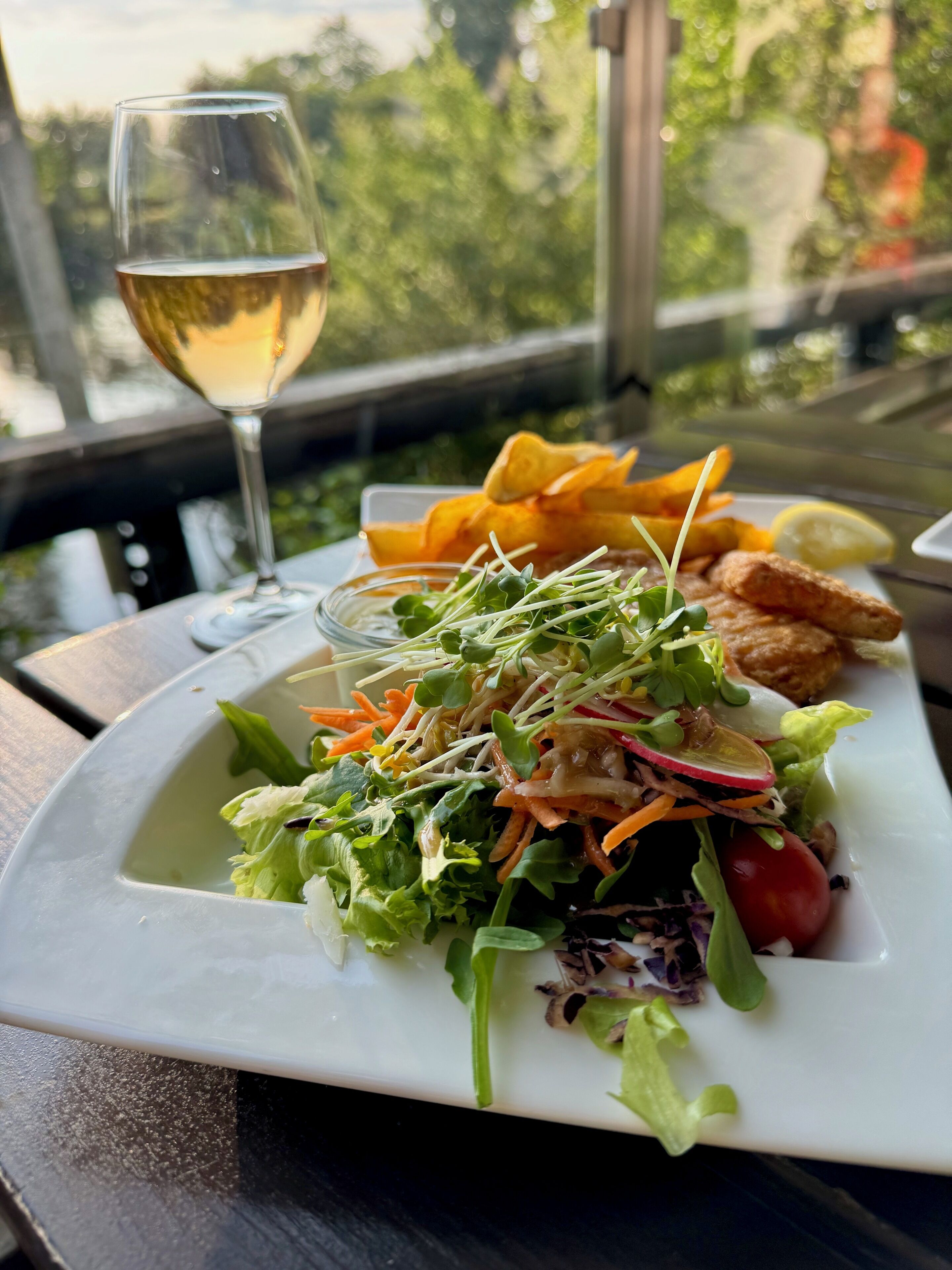 Un plato de comida con ensalada, papas fritas y una copa de vino blanco sobre una mesa con vista a árboles.