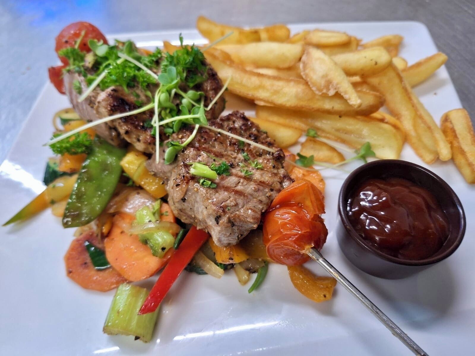 A plate of food with grilled meat, fries, and vegetables, and a small bowl of sauce.