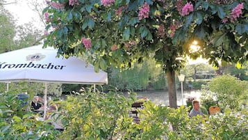 An outdoor seating area with a white umbrella labeled "Krombacher," surrounded by lush greenery and trees.