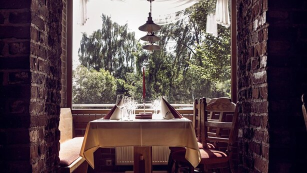 A dining area with a table set for two, a brick wall, and a window with a view of trees. - Altes Eishaus, Hotel & Restaurant (Giessen)