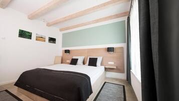 A bedroom with a bed, a headboard, a nightstand, a wall-mounted lamp, a window with curtains, and a small rug.