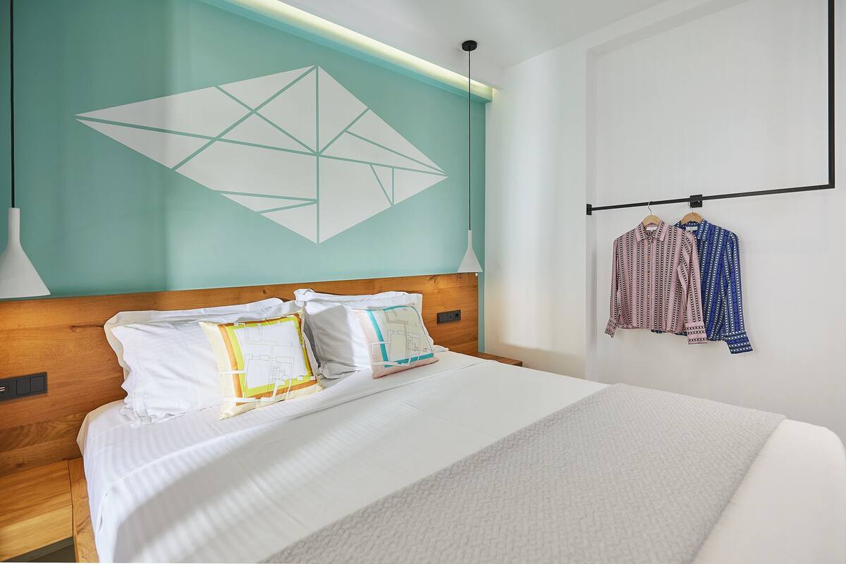 A neatly made bed with white linens, a geometric wall design, and a clothing rack with patterned shirts.
