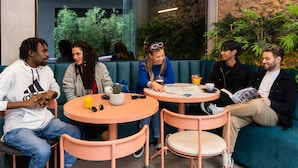 A group of people seated at a round table with a menu, surrounded by teal seating and a glass wall with greenery. - Mosaikon - Hostel (Athens)