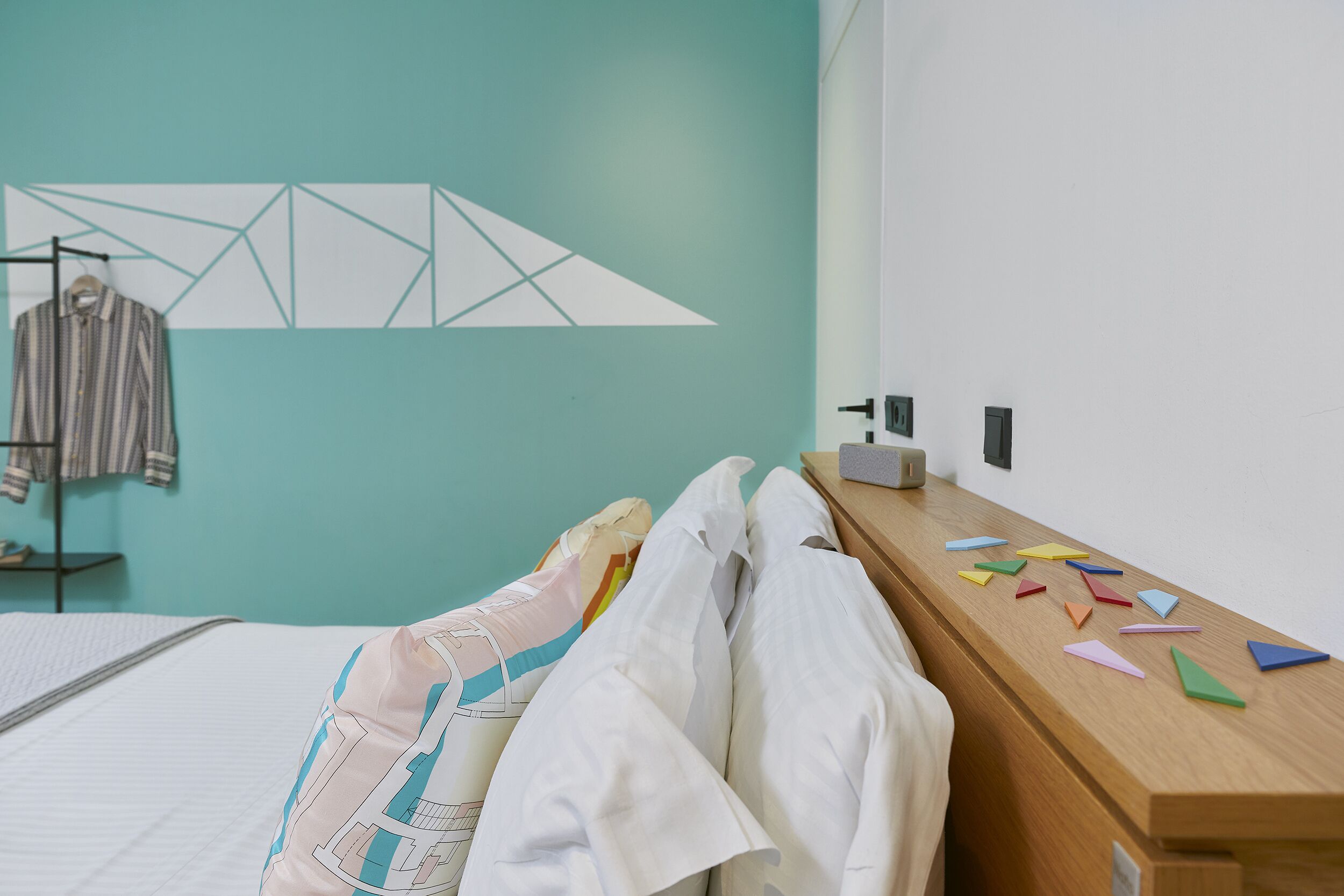 Quarto de hotel com parede verde-água, cama com roupa de cama branca, mesa de cabeceira de madeira com formas geométricas coloridas e um cabideiro com roupas.