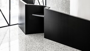A modern reception area with a geometric glass partition, a black reception desk, and a speckled textured wall. - Mosaikon - Hostel (Athens)
