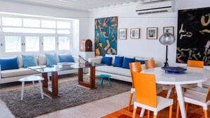 A modern living room with a white sofa, blue and white cushions, a glass coffee table, and a round dining table with orange chairs.