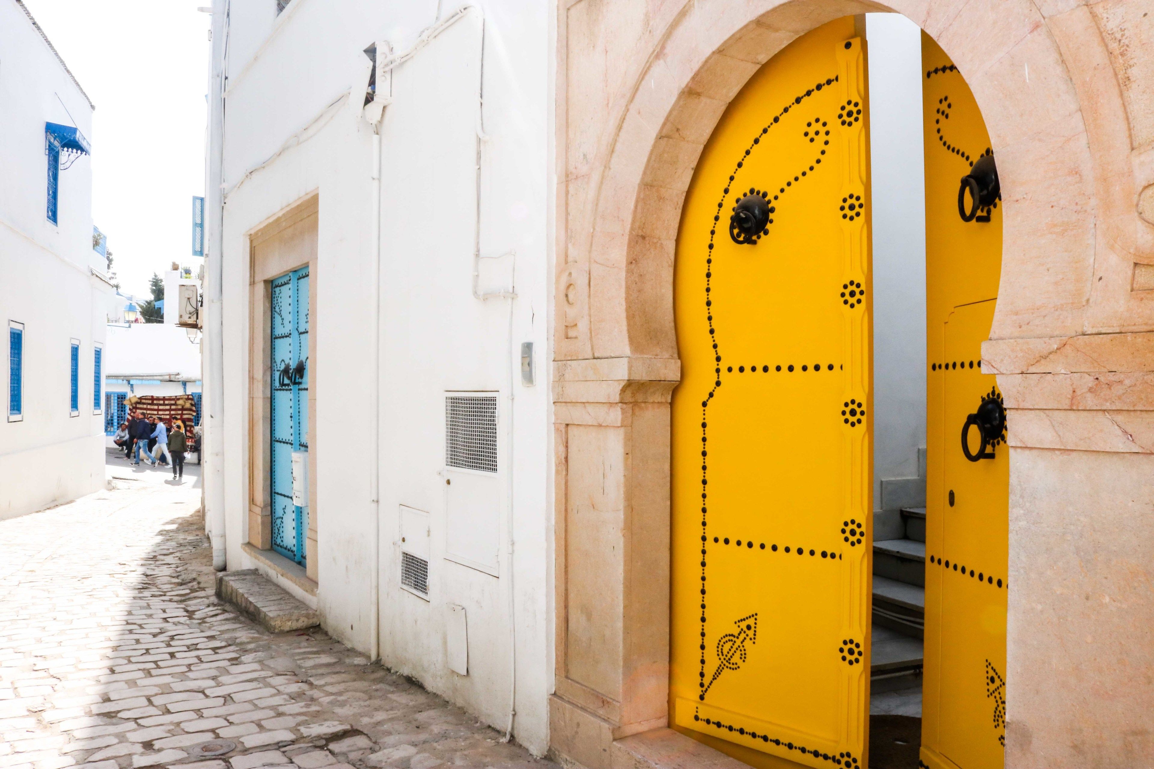A narrow cobblestone street with a white building featuring a blue door and a yellow door with decorative patterns.