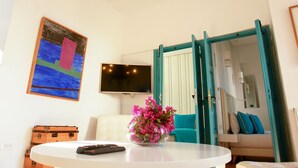 Una habitación con una mesa blanca, un jarrón de flores rosadas, un televisor en la pared y una puerta con marco color turquesa.