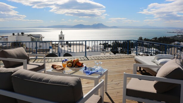 Una terraza con vistas al mar, una mesa con frutas y copas, y sillones de descanso.