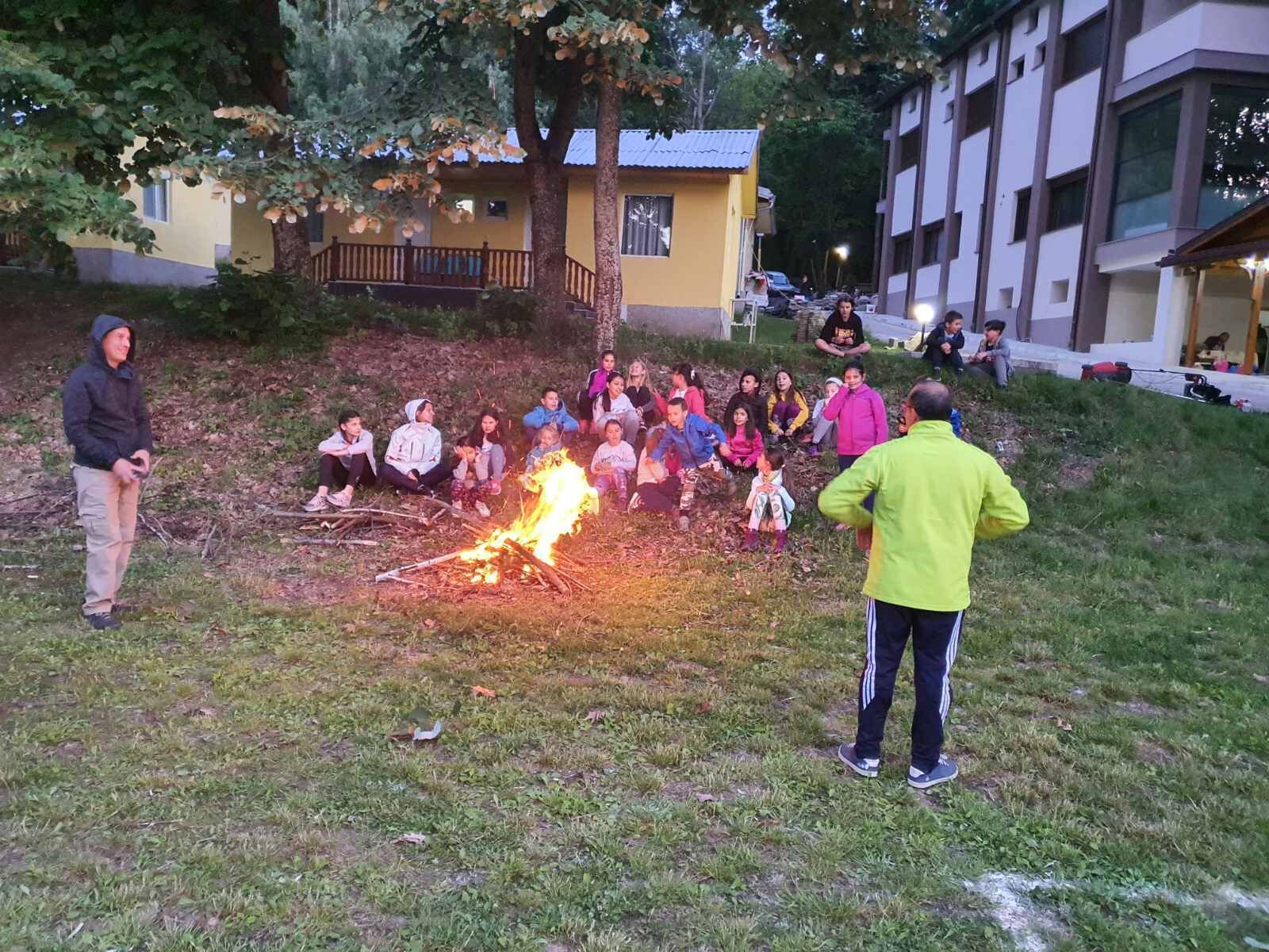 A group of people gathered around a campfire in an outdoor setting with buildings in the background.