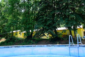 Una piscina con superficie de agua azul cristalina, rodeada de frondosos árboles verdes y un edificio amarillo al fondo.