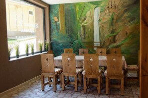 A dining area with wooden chairs and a table, a mural on the wall, and a window with plants. - Bungalows Kamena (Petrich)