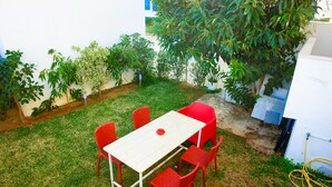 A small outdoor patio with a white table and red chairs, surrounded by green grass and various plants. - The View (La Marsa)