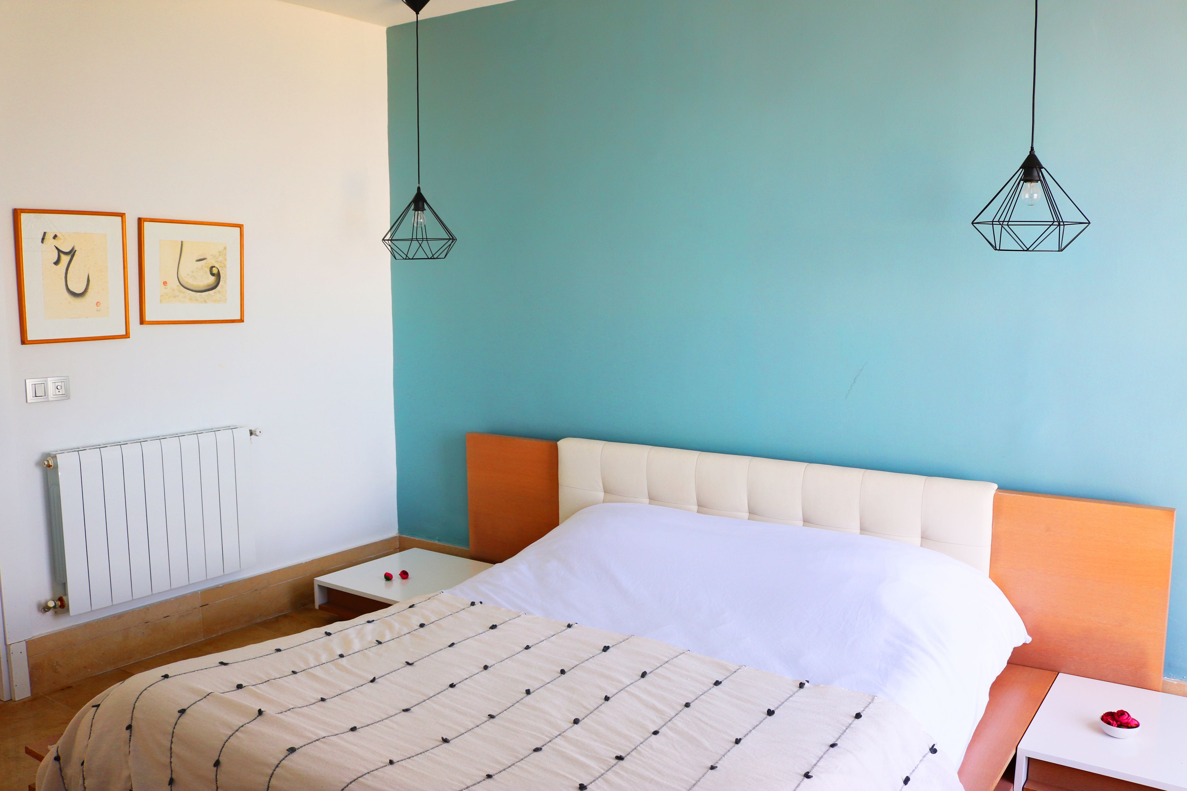 A bedroom with a bed, a bedside table, a nightstand, a radiator, and two wall-mounted light fixtures.