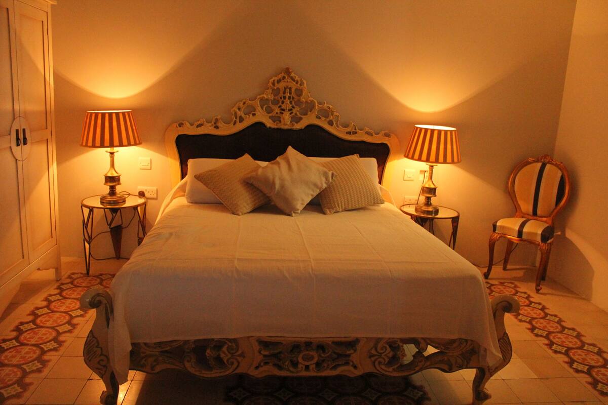 A neatly made bed with a decorative headboard, two bedside lamps, and a chair with a mirror.