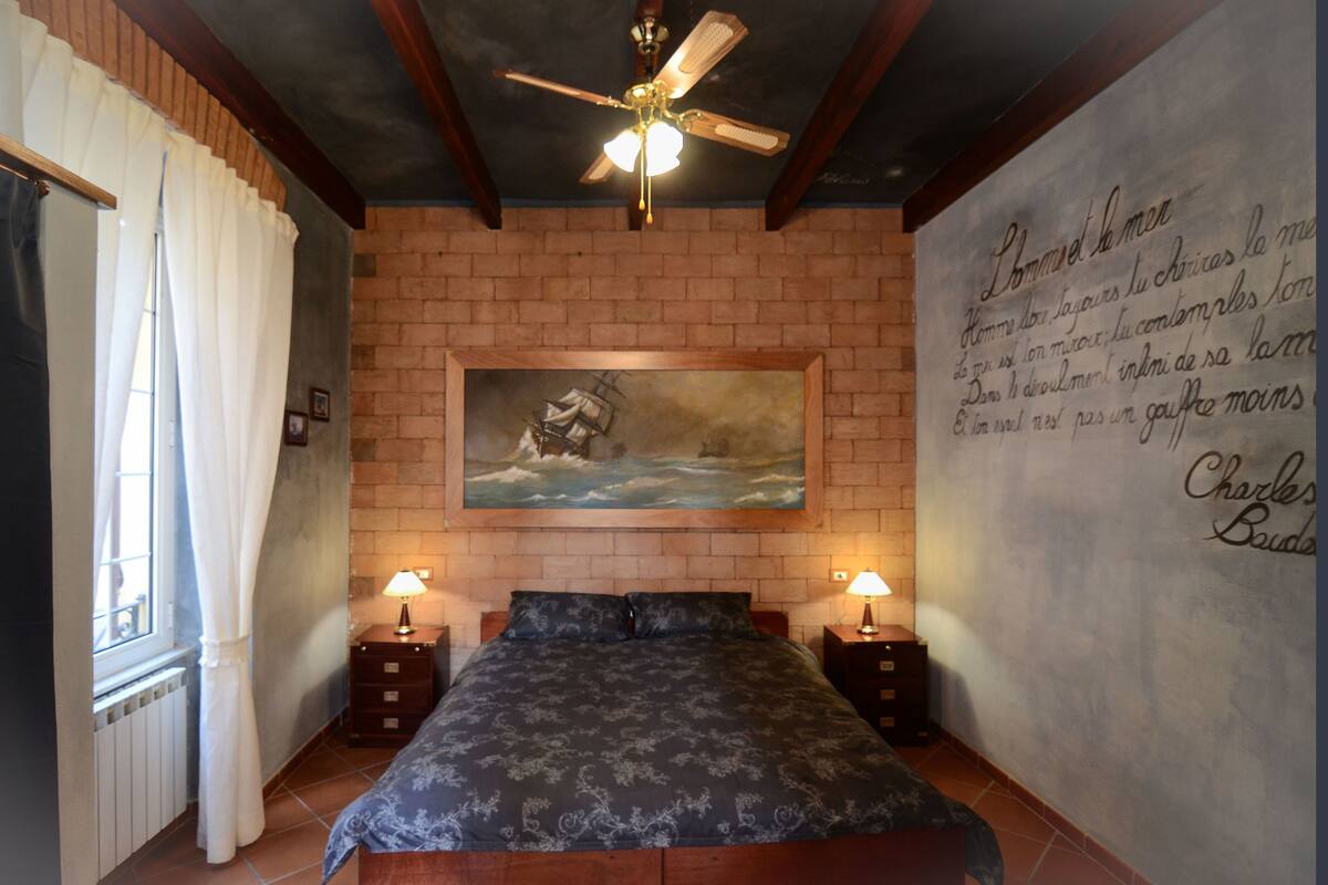Une chambre avec un mur en briques, un lit, deux tables de chevet avec des lampes, un ventilateur de plafond et un tableau représentant un navire accroché au mur.