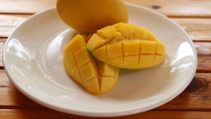 Um prato branco com um mango inteiro e várias fatias de mango sobre uma mesa de madeira.