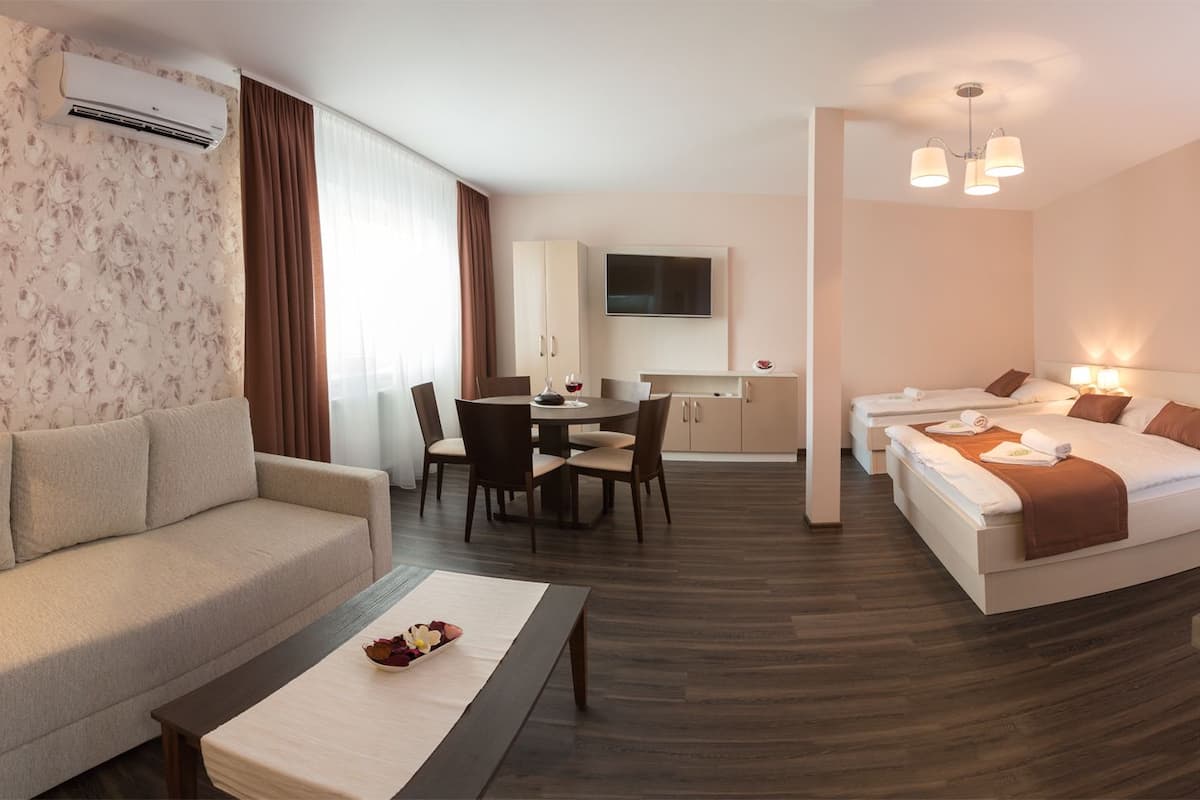 A hotel room with a sofa, a dining table with chairs, a bed with two pillows, a TV, a kitchen area with a sink and stove, and a window with curtains.