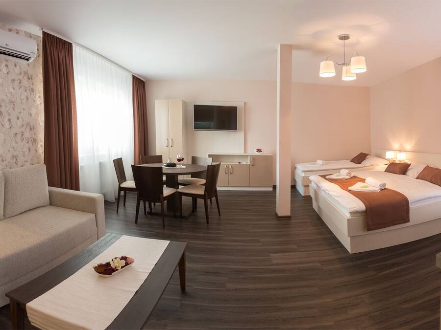 A hotel room with a sofa, a dining table with chairs, a bed with two pillows, a TV, a kitchen area with a sink and stove, and a window with curtains.