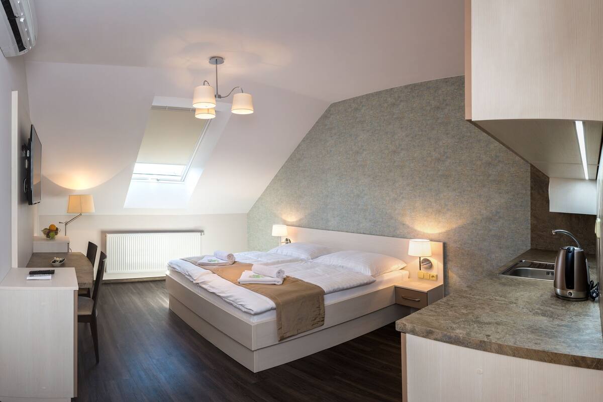A modern bedroom with a loft-style design, featuring a bed, bedside tables, a desk, and a kitchenette.