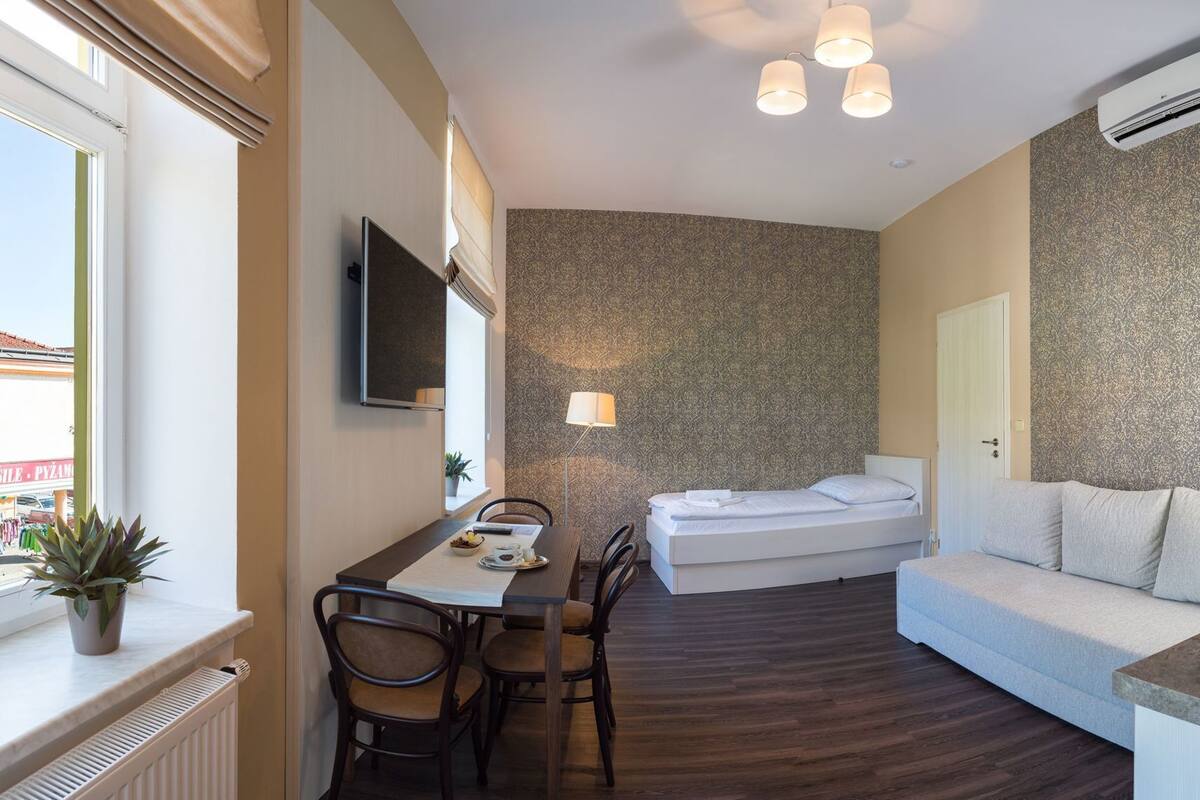 A compact hotel room with a bed, a sofa, a dining area with chairs, a TV, and an air conditioning unit.