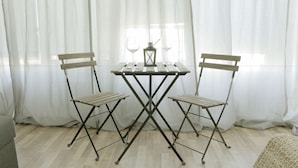 A room with a wooden table and two foldable chairs, a small table lamp, and sheer curtains. - Cozy apARTment (Sofia)