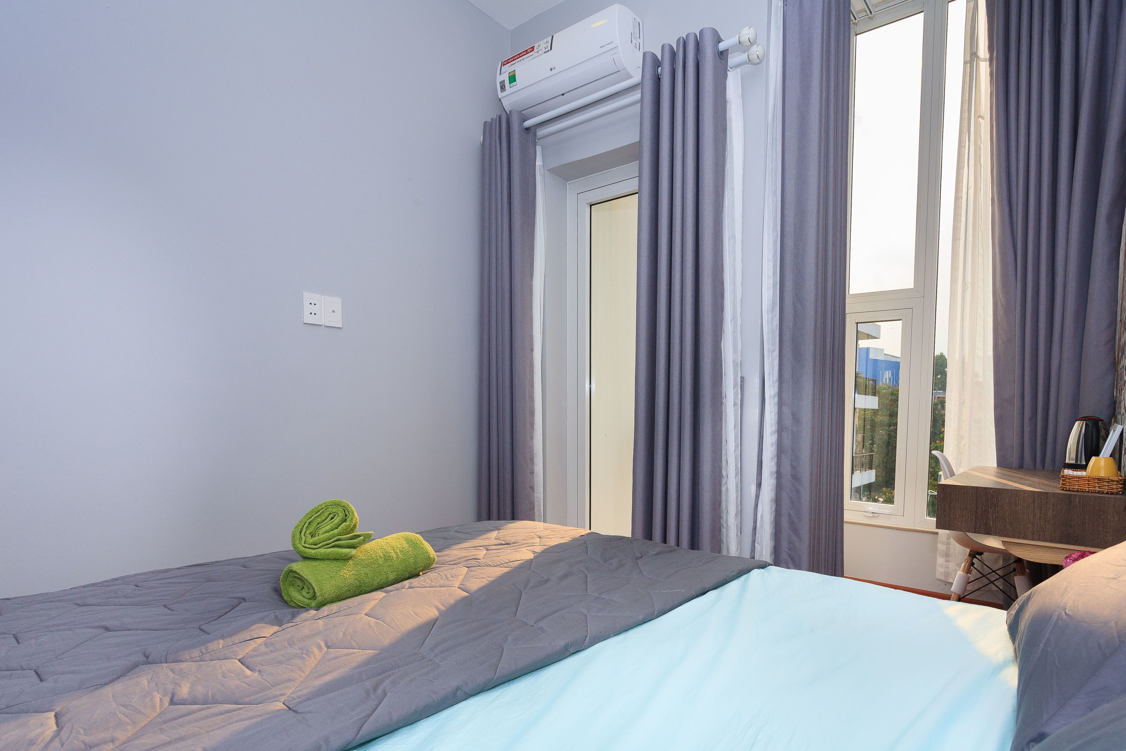 Une chambre à coucher avec un lit, une fenêtre avec des rideaux, un climatiseur et une petite table avec une lampe.