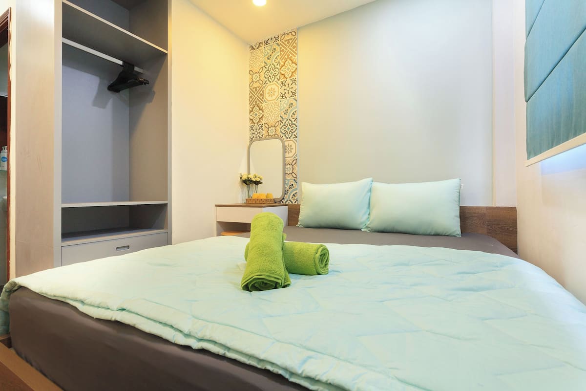 Een net opgemaakt bed met een lichtblauwe spreideken en twee groene handdoeken, een nachtkastje met een bloevas, een spiegel met een versierde lijst, en een kledingkast met een hangroede.
