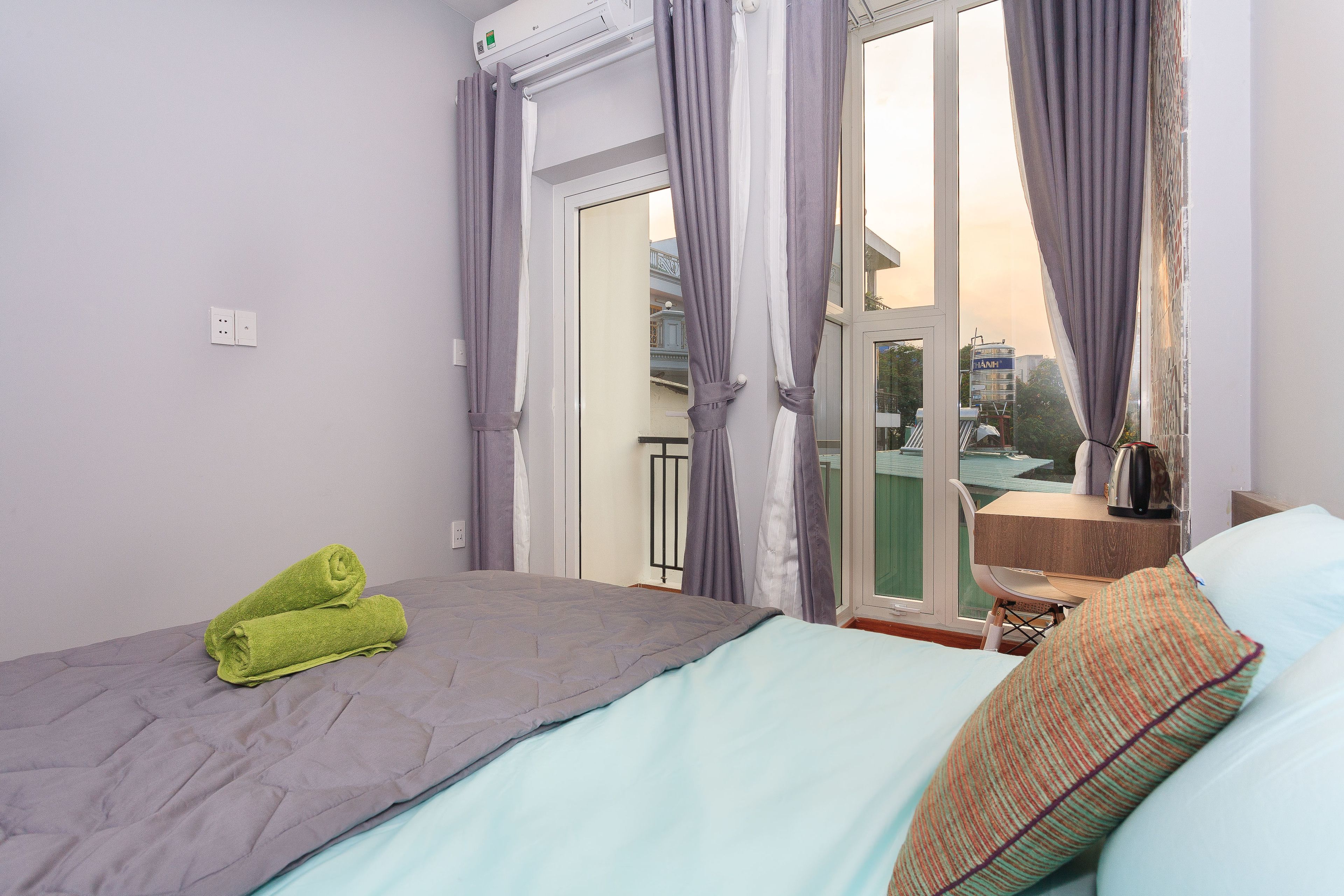 A bedroom with a bed, a window with curtains, a view of the outside, and a small table with a chair.