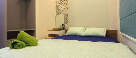 Una cama bien hecha con edredón azul y blanco, una toalla verde y una mesita de noche con hervidor y taza.