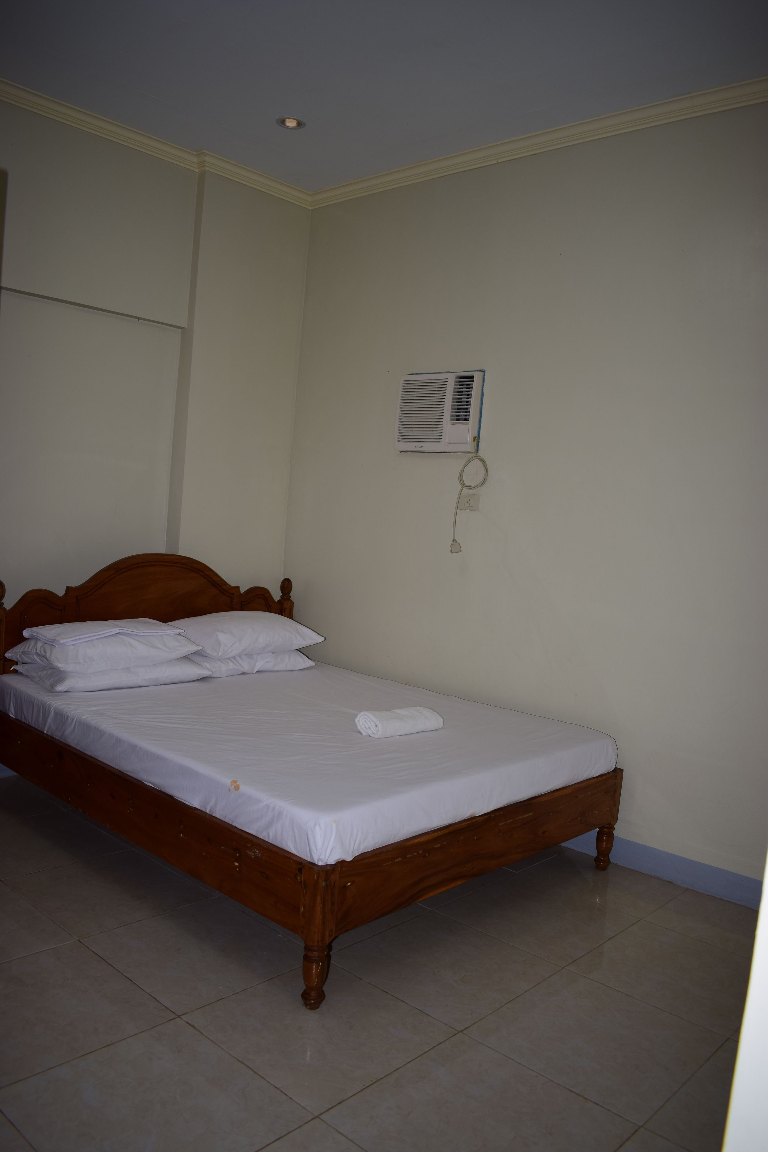 Uma cama de solteiro com cabeceira de madeira e lençóis brancos em quarto com paredes lisas e chão de azulejos.