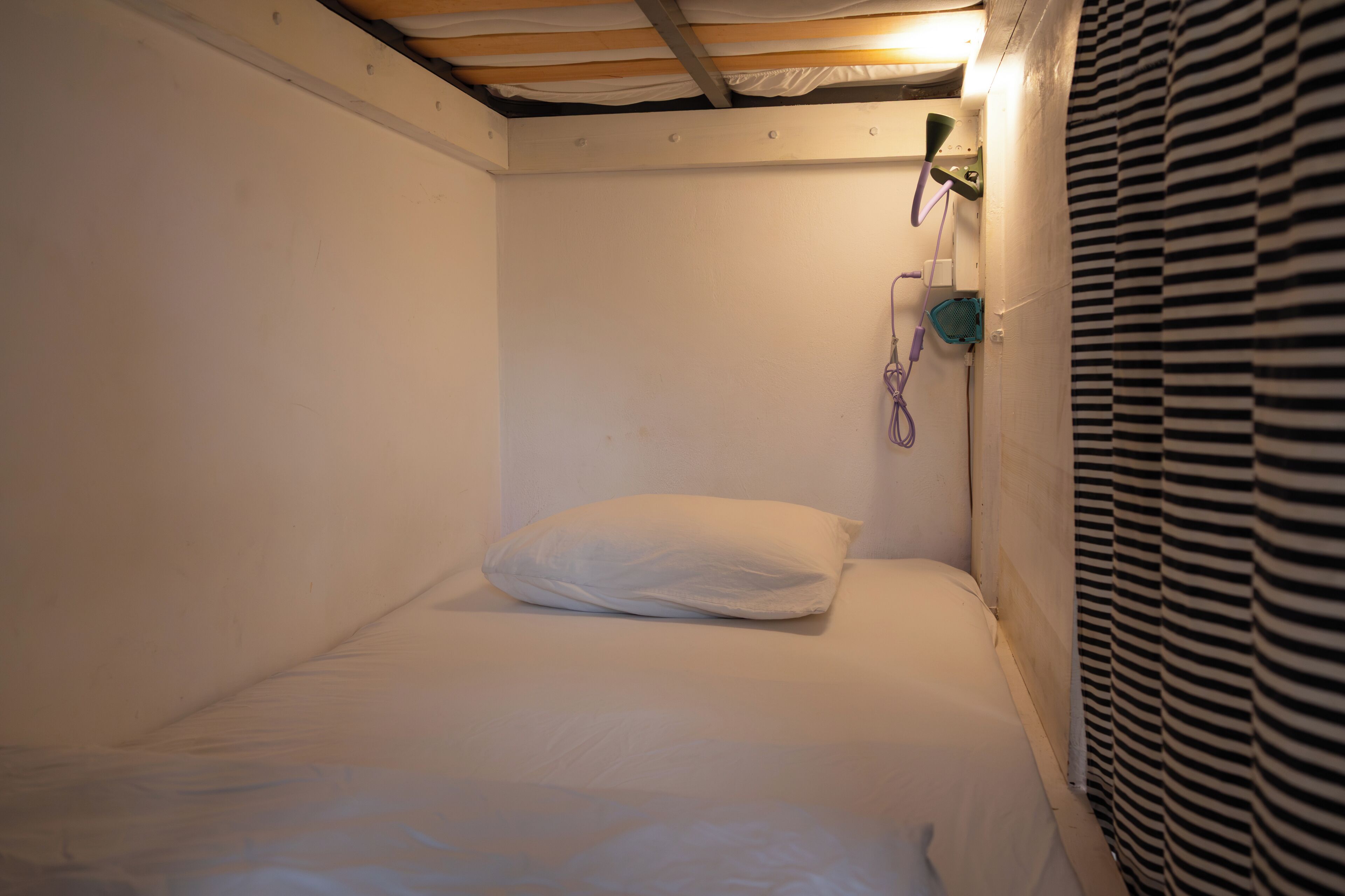 Habitación pequeña con una cama individual, colchón y almohada blancos, techo de madera y cortina a rayas.