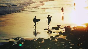 Escena de playa con gente caminando por la orilla, algunos llevando tablas de surf.