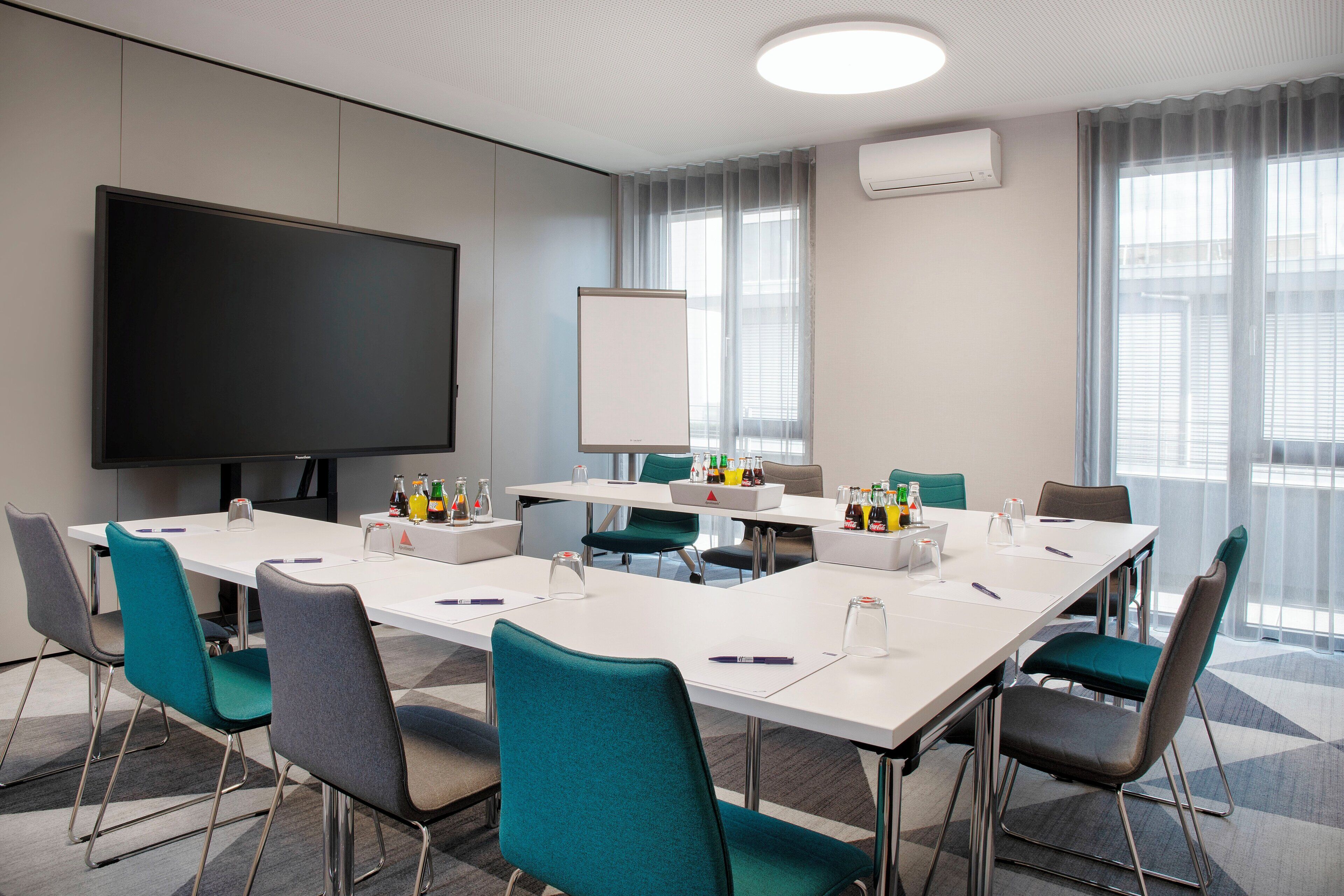 Une salle de conférence équipée d’un grand téléviseur à écran plat, de tables blanches, de chaises de couleur turquoise et d’un tableau blanc.
