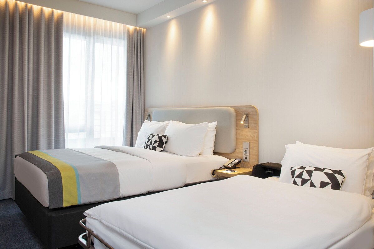 Une chambre d’hôtel avec deux lits, une table de chevet avec un téléphone et une fenêtre avec des rideaux.