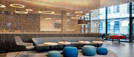 Un interior moderno con una zona de estar que incluye sofás y mesas redondas, una pared decorativa con luces colgantes y grandes ventanales que ofrecen vistas al exterior.