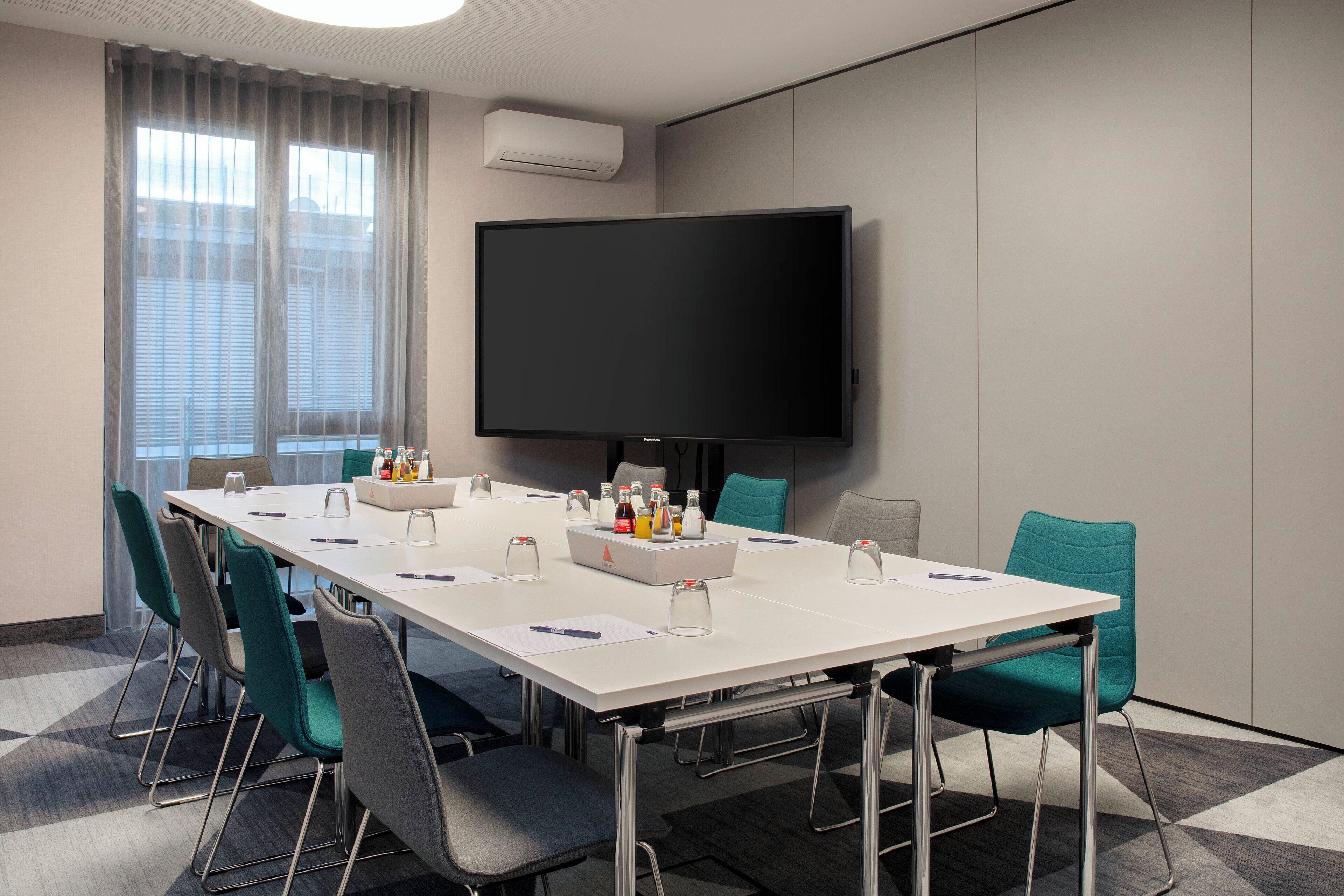 A conference room with a long table, chairs, a flat-screen TV, and a window with blinds.