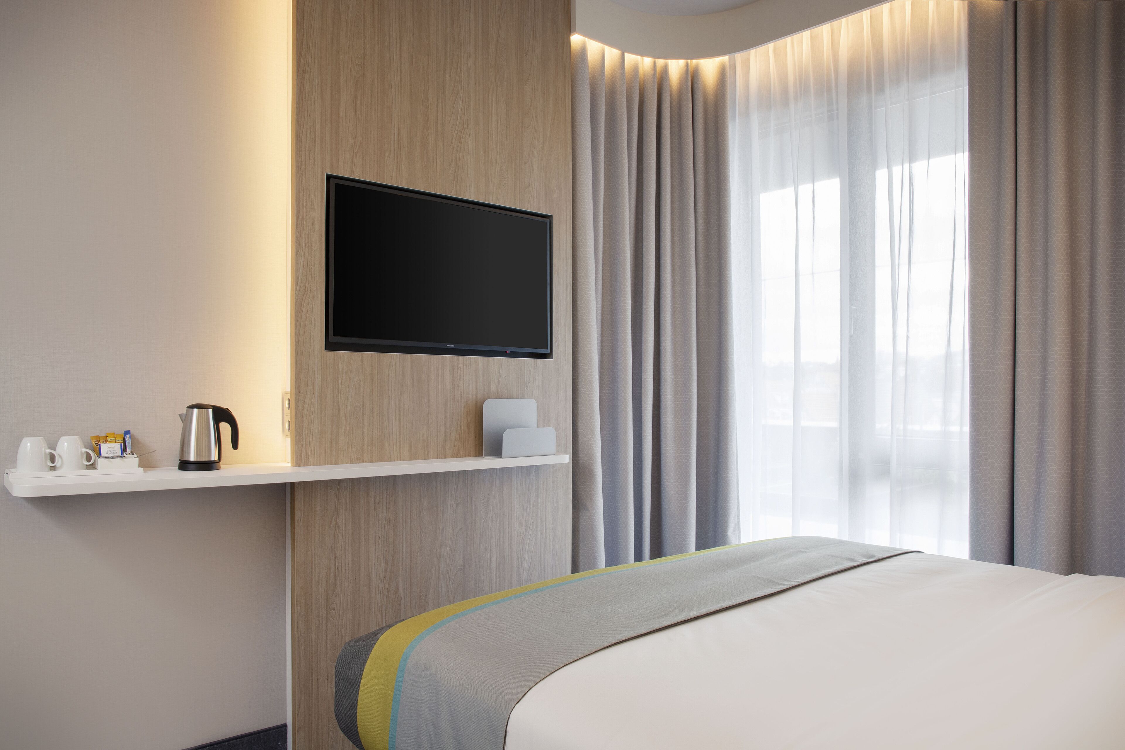 A hotel room with a bed, a mounted television, a shelf with a kettle and cups, and a window with curtains.