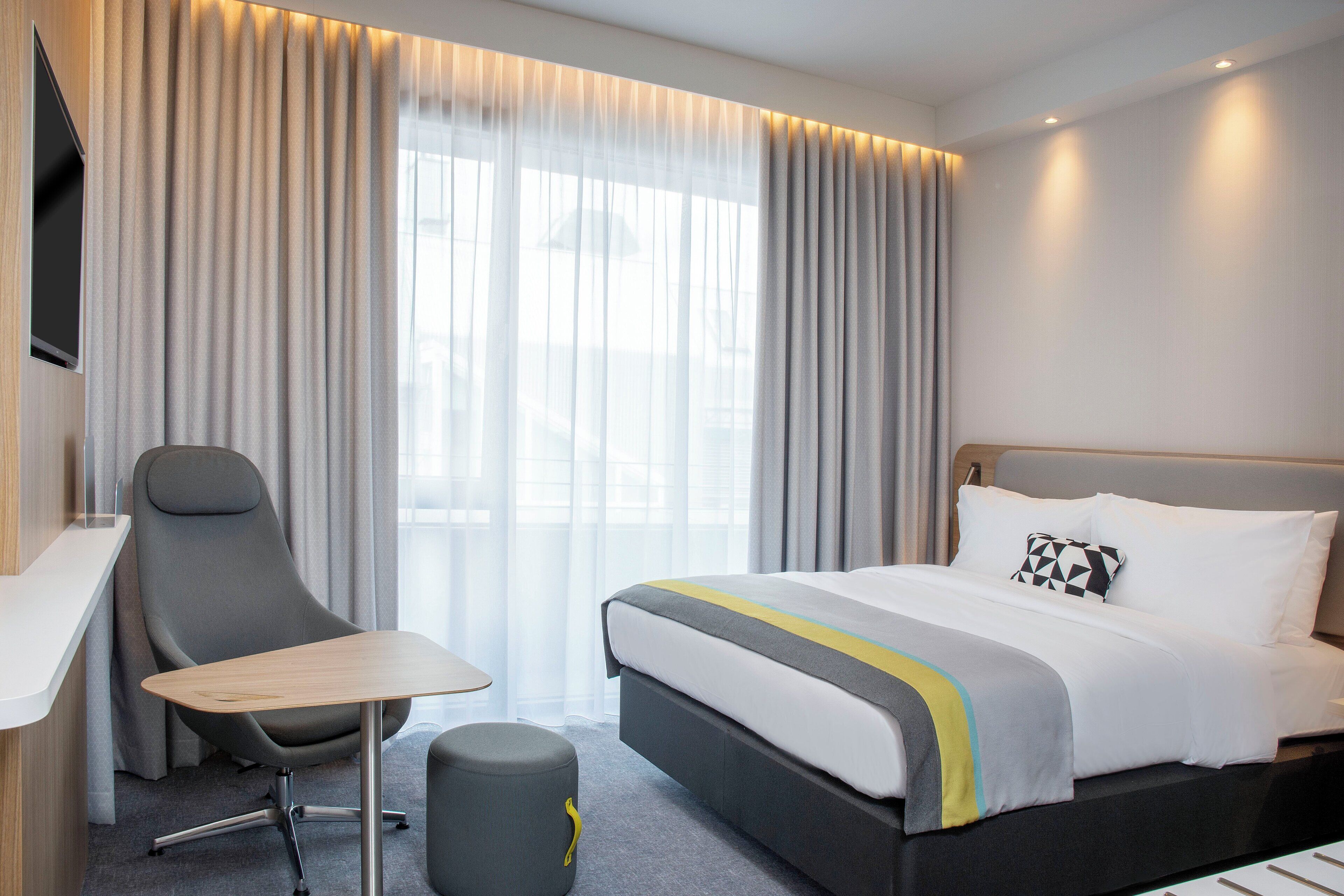 Habitación de hotel con cama, escritorio, silla, televisor y ventana con cortinas.