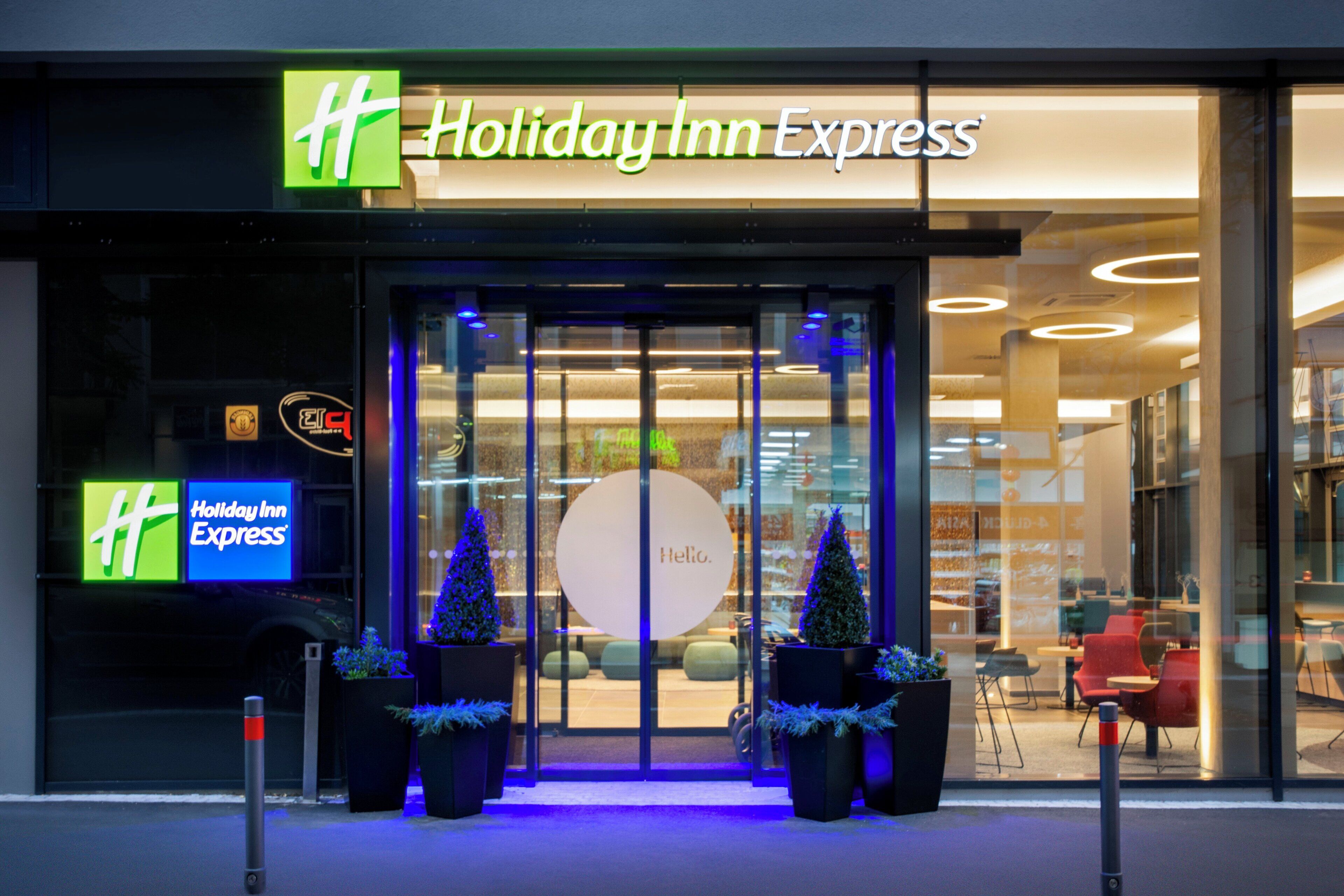 Ingången till ett Holiday Inn Express-hotell med en glasdörr, två julgranar i krukor och en skylt med texten ”Hej”.