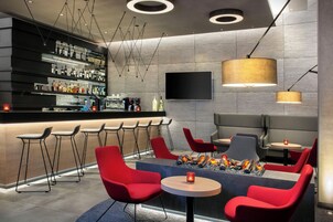 Zona de bar moderna con una barra larga, varios taburetes y un área de estar con mesas redondas y sillas rojas.